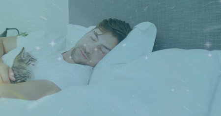 Lying adult man wearing white t-shirt, cradling tabby kitten on bed in bedroom, with bedside lamp. Comfort, relaxation, intimacy, tranquility, domestic, cozy, wellnessの写真素材