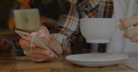 Holding smartphone and ceramic coffee cup, man sitting at coffee shop table, with saucer and laptop....