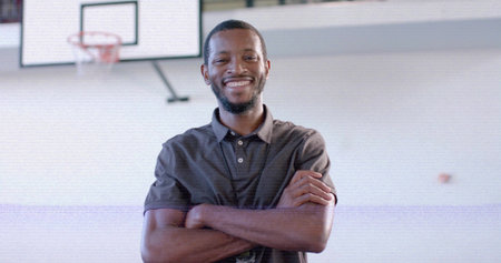 Crossing arms coach wearing dark grey polo shirt inside basketball gym, with hoop and backboard. Athletic, sport, teamwork, training, motivation, fitness, interiorの写真素材