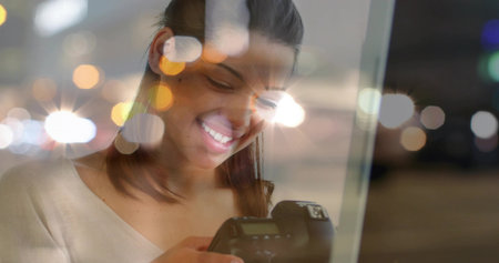 Smiling woman examining DSLR camera display inside apartment at night, with bokeh reflections. Portrait, nightlife, urban, bokeh, reflection, creative, introspectionの写真素材