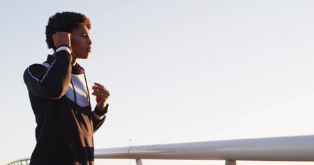 Adjusting earbuds runner gazing left on pedestrian bridge at dawn, with metal railing, copy space....