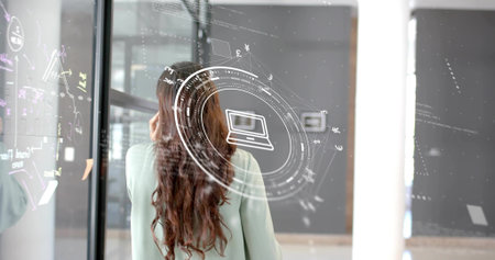 Studying Indian woman viewing digital schematics on glass in office corridor, with laptop icon. Technology, innovation, futurism, corporate, data, touchscreen, digitalの写真素材