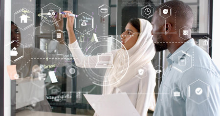 Collaborating coworkers in hijab and shirt annotating glass board at office, with holographic icons....