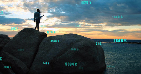 Standing hooded woman on boulders at sunset, with smartphone showing AR currency values, copy space. Silhouette, adventure, digital, skyline, tranquility, exploration, innovationの写真素材