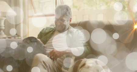 Middle-aged man wearing eyeglasses, olive shirt petting two dogs on sofa in living room with lamp. Relaxation, companionship, cozy, lifestyle, natural light, domestic, comfortの写真素材