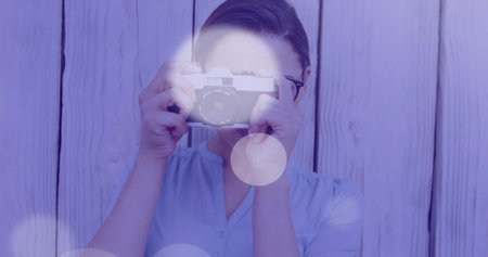 Standing female photographer holding film camera against wooden plank wall under soft lighting....