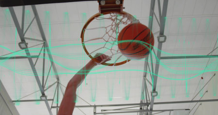 Reaching male athlete's jersey-clad arm scoring basketball through hoop and net in gym with trusses....