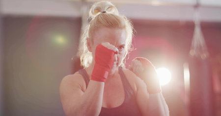 Boxer wearing sports tank top maintaining boxing stance in gym, with red wraps and punching bags. Fitness, strength, determination, training, athletic, empowerment, dedicationの写真素材