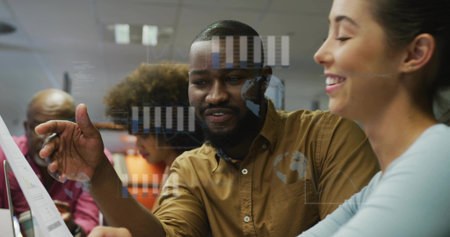Gesturing man in brown shirt pointing at printed charts in office, with digital graphs and globe. Collaboration, teamwork, analysis, data, business, technology, professionalの写真素材