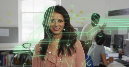 Smiling mid adult businesswoman in office, with coworkers working at monitors and circuit overlay. Collaboration, teamwork, innovation, modernity, professionalism, diversity, productivityの写真素材