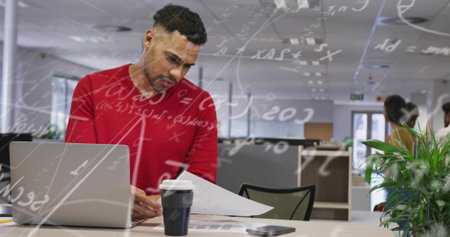 Reviewing man in red sweater analyzing printed charts at open plan office, with laptop smartphone....