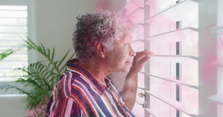Senior woman wearing striped top peering through blinds at home, with plant, earrings, copy space. Retirement, relaxation, tranquility, domestic, sunlight, natural light, peacefulの写真素材