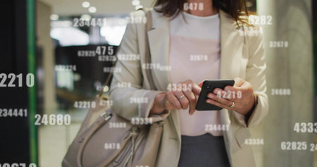 Tapping professional woman wearing blazer holding smartphone in lobby, with floating numeric data. Corporate, professionalism, technology, modern, businesswoman, digital, dataの写真素材