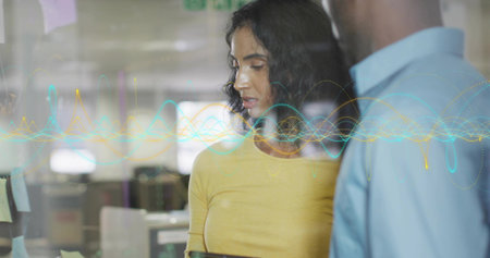 Reviewing woman wearing yellow top holding tablet beside partition in office, with notes, waveforms....