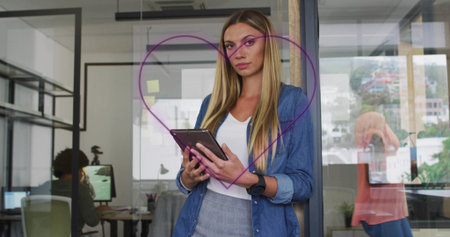 Standing blonde woman holding tablet inside glass meeting room, with purple heart drawing. Collaboration, teamwork, professionalism, modern, corporate, communication, innovationの写真素材