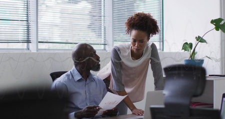 Collaborating senior man and woman reviewing report on laptop at desk in office, with potted plant. Collaboration, professionalism, productivity, teamwork, modern, workspace, communicationの写真素材