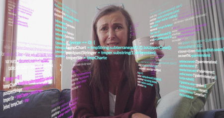 Holding ceramic mug, woman wearing cardigan frowning on gray sofa at home, with code overlay....