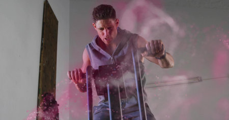 Working out man in vest and shorts gripping elliptical display, handles in gym swirling pink powder....