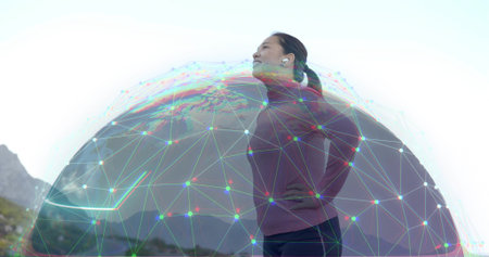 Standing Asian woman wearing activewear at mountain vista, with wireless earbuds and digital sphere....
