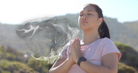Adult Asian woman praying on sunlit hillside, with pink Tshirt and fitness tracker, copy space....