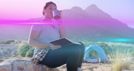 Woman camper holding book, lifting metal mug, sitting on rock in canyon campsite, with blue tent. Adventure, outdoor, wilderness, exploration, rugged, relaxation, naturalの写真素材