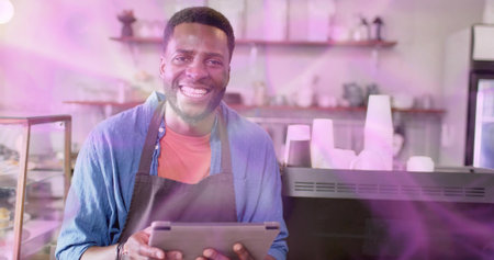 Smiling male barista wearing red shirt and dark apron, holding tablet at cafe, with coffee machine....