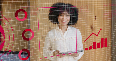 Presenting businesswoman in white blouse using tablet with grid overlay and red charts in office. Technology, data analytics, professional, modern, digital, corporate, innovationの写真素材