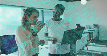 Standing women wearing office attire collaborating at open-plan office desk with laptop and mug. Collaboration, teamwork, professional, workspace, daylight, modern, productivityの写真素材