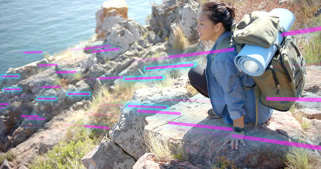Crouching Asian hiker viewing rugged cliffside inlet, carrying backpack with rolled mat, copy space....