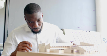 Examining architect wearing white shirt studying scale model in office, with pencil and chart board....
