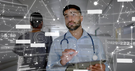 Interacting male doctor scrolling data on tablet in medical hallway, with digital overlay graphics. Healthcare, digital, innovation, professionalism, technology, teamwork, hospitalの写真素材