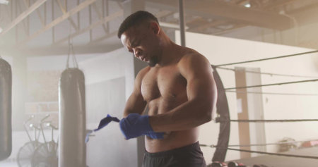 Wrapping hands, muscular boxer tightening blue hand wraps in boxing ring, with punching bag. Strength, fitness, training, dynamic, athleticの写真素材