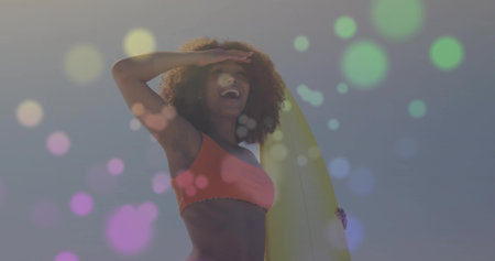 Smiling adult female surfer shielding eyes at sandy beach, showcasing yellow surfboard and bikini. Adventure, leisure, vibrant, energetic, outdoors, lifestyle, positivityの写真素材