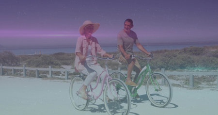Riding couple pedaling pastel cruisers along sandy coastal path, with sunhat, white fence and dunes. Scenic, leisure, vacation, adventure, outdoor, tranquility, navigationの写真素材