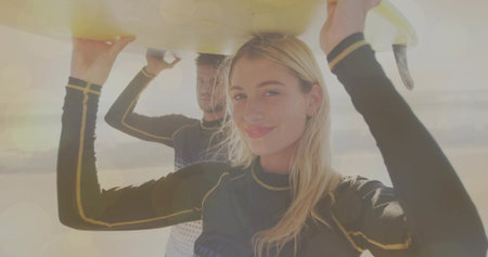 Holding yellow surfboard, woman and Hispanic man wearing wetsuits walking on sandy beach at sunset....