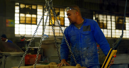 Operating chain hoist lifting casting at plant, worker in coverall using yellow remote, copy space....