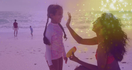 Kneeling mother holding sunscreen bottle applying sunscreen to daughter on sandy beach, with waves. Family, beach, outdoor, summer, recreation, leisure, sunnyの写真素材