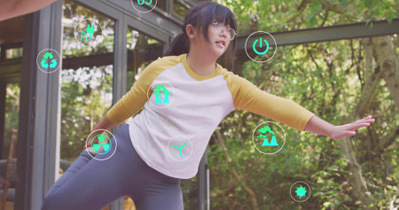 Balancing Chinese child wearing raglan shirt and leggings at glass pavilion with floating eco icons. Environment, sustainability, eco-friendly, education, childhood, innovation, natureの写真素材