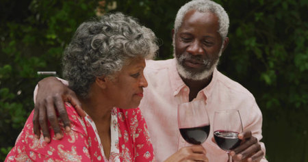 Toasting senior couple raising stemmed red wine glasses on garden patio, with dense green foliage....