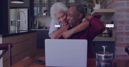 Hugging senior woman wrapping arms around seated man at kitchen table, with laptop French press. Affection, companionship, warmth, modern, cozy, lifestyle, intimacyの写真素材
