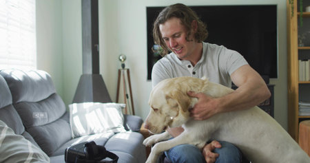 Man holding Labrador retriever and sitting on grey sofa in living room, with VR headset. Coziness, relaxation, modern, domestic, comfort, technology, home interiorの写真素材