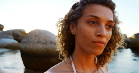 Standing woman wearing thin strap swimsuit top and chain necklace at shore at sunset, with boulders. Scenic, natural, serene, twilight, coastal, meditative, minimalistの写真素材