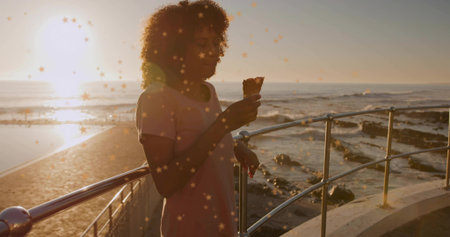 Woman wearing pale pink dress leaning against railing during coastal sunset, holding ice cream cone....