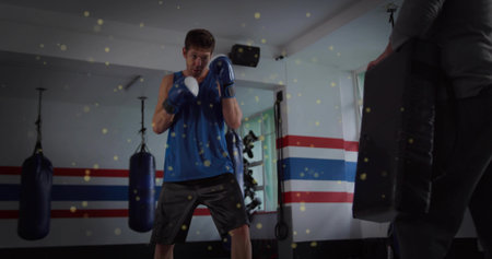 Practicing boxer wearing blue top and gloves throwing punches in gym, with trainer holding pad....