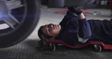 Lying mechanic inspecting vehicle underside in repair garage, with red creeper and work gloves. Automotive, repair, engineering, industrial, tools, maintenance, craftsmanshipの写真素材