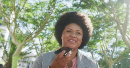 Speaking woman wearing gray blazer, pink blouse holding smartphone near mouth in park, green trees....