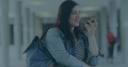 Speaking woman wearing denim jacket using smartphone microphone in transit hub, with blue backpack....