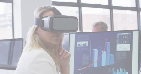 Resting female wearing VR headset, white blazer at office desk, with monitors showing upward trend....