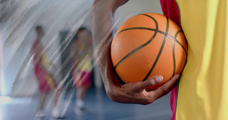 Basketball player holding orange ball at torso on gym court with teammates in jerseys, copy space. Sports, teamwork, athleticism, endurance, dynamic, vibrant, competitionの写真素材