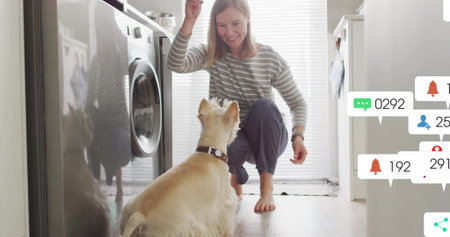 Kneeling woman holding treat above small collared dog in laundry area with washer and notifications. Pet, domestic, cozy, modern, lifestyle, interaction, minimalistの写真素材
